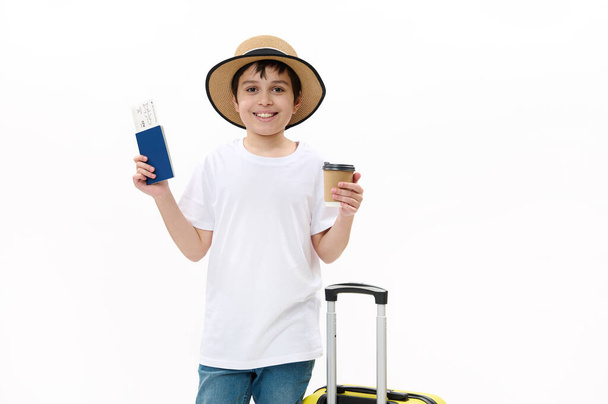 Joyeux garçon touriste adolescent en jeans bleus, t-shirt décontracté et chapeau de paille avec sac de voyage, tient une boisson chaude à emporter dans une tasse en carton jetable et une carte d'embarquement, isolée sur fond blanc - Photo, image