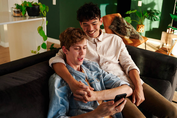 Happy young gay couple smiling while looking at mobile phone and holding hands on sofa at home - 写真・画像