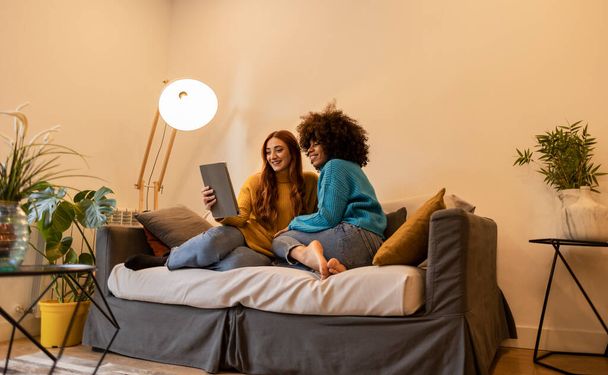 Novias lesbianas multirraciales viendo las redes sociales en la tableta en el sofá. Feliz pareja de mujeres en la automatización del hogar inteligente. - Foto, imagen