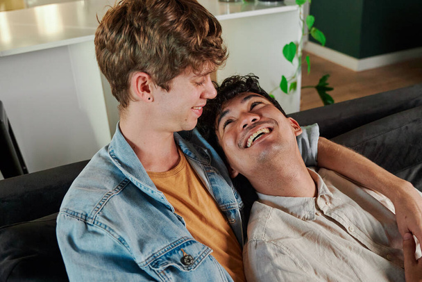 Happy young gay couple smiling while relaxing face to face on sofa in living room at home - Zdjęcie, obraz