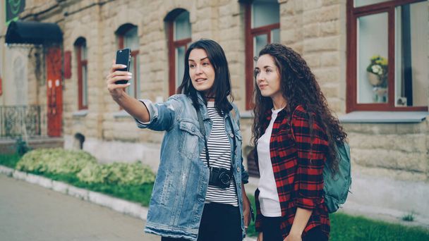 Emotional young women happy travelers are making online video call using smartphone holding device and talking showing historical building behind them expressing excitement. - Zdjęcie, obraz
