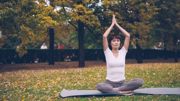 Beautiful girl is concentrated on yoga practice sitting on mat in park in lotus position and bending sideways raising arm. Healthy lifestyle, sports and recreation concept. - Foto, afbeelding