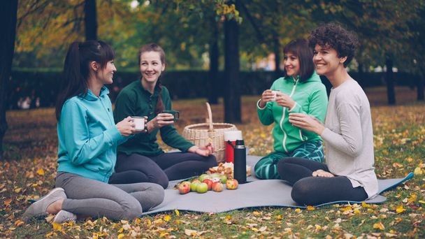 Happy girls are having picnic in park sitting on yoga mats and eating after outdoor practice in autumn, girls are talking and laughing. Communication, people and food concept. - Zdjęcie, obraz