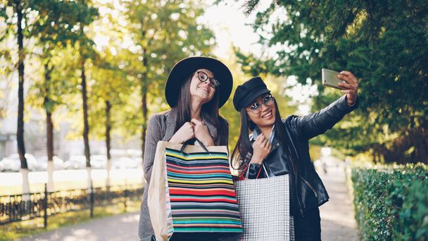 Happy young women friends are taking selfie with colorful paper bags standing outdoors on sidewalk and posing. Girls are using smartphone and wearing casual clothing. - Foto, afbeelding