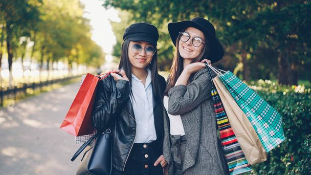 Portrait of beautiful young women shoppers standing in the street holding paper bags and smiling looking at camera. Youth lifestyle, friendship and consumerism concept. - 写真・画像