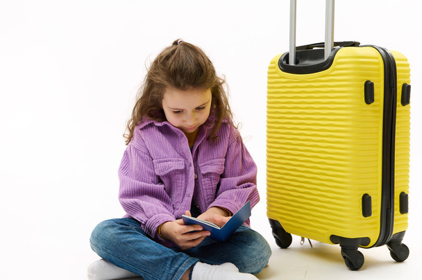 Adorable little child, traveler baby girl wearing blue jeans and purple shirt, sitting near a yellow suitcase with boarding pass in her hands, going for weekend getaway, isolated over white background - Фото, зображення