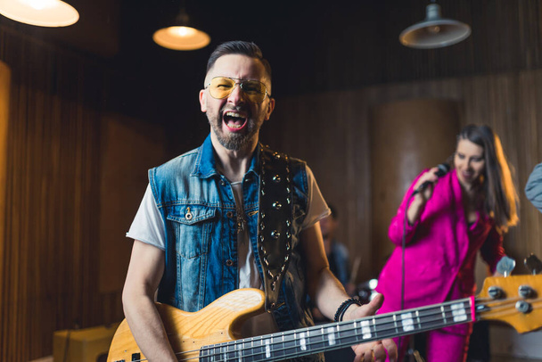 Kaukasischer Mann in Jeans, ärmelloser Weste, E-Gitarre in der Hand und schreiend. Indoor-Schuss. Band im Tonstudio. Hochwertiges Foto - Foto, Bild