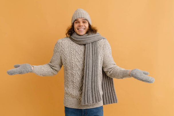 Young handsome man with long hair in sweater and winter hat, scarf and mittens spreading arms to sides over isolated brown background - Zdjęcie, obraz