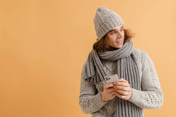 Young handsome man with long hair in winter hat and scarf holding phone and looking rightward over isolated brown background - Foto, imagen