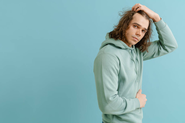 Young handsome man with long hair in hoodie posing with his right side over isolated blue background - Photo, Image