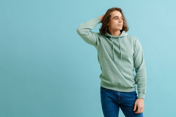 Young handsome man in hoodie touching his long hair and looking rightward over isolated blue background - Foto, Imagem