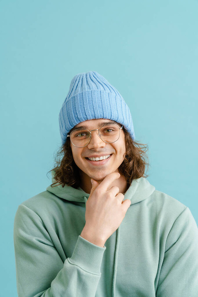 Young handsome man with long hair in a hoodie, glasses and winter hat touching his chin and standing over isolated blue background - Foto, Imagen