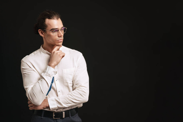 Young handsome man in shirt and glasses touchung his chin and looking rightward over isolated black background - Φωτογραφία, εικόνα