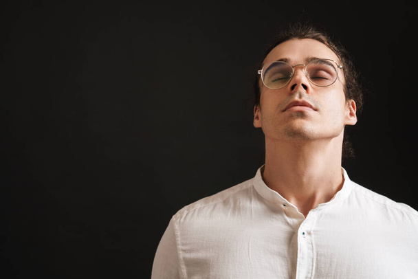 young handsome man in shirt and glasses with raised head and closed eyes over isolated black background - Foto, Imagen