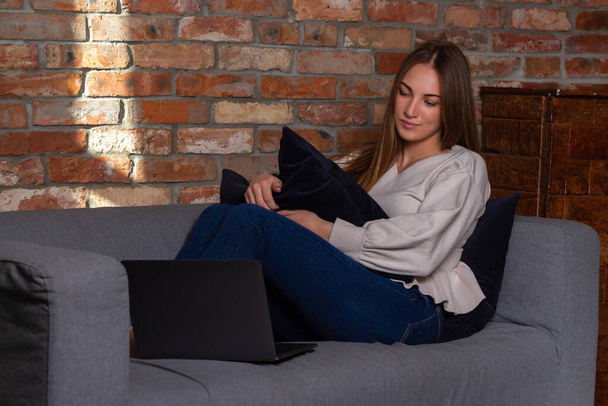 Smiling woman in a white sweater sitting on the couch, holding a pillow and looking at a laptop - Foto, Imagen