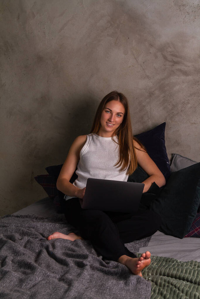 Smiling woman in a white top sitting on a bed with a laptop in her lap and looking into the camera - Photo, image