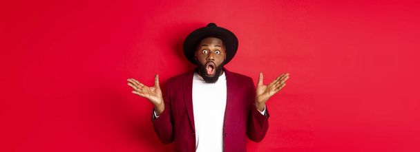 Fashion and party concept. Impressed Black man in classy outfit staring with complete disbelief at camera, gasping and looking surprised, standing over red background. - Photo, image