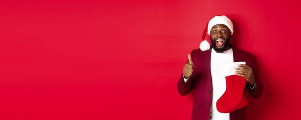 Emocionado hombre negro mostrando el pulgar hacia arriba en la aprobación, la celebración de calcetines de Navidad con regalos de vacaciones, sonriendo sorprendido, de pie sobre el fondo rojo. - Foto, imagen
