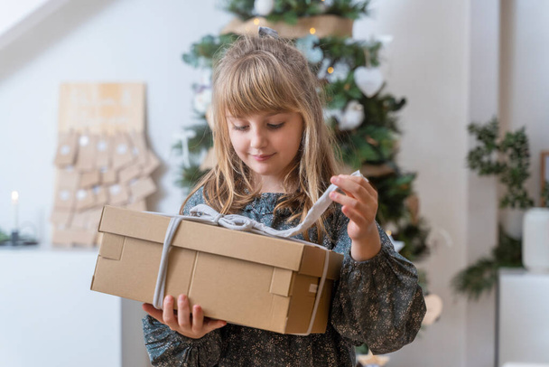 Retrato interior original de una niña abriendo un regalo cerca del árbol de Navidad en casa - Foto, imagen