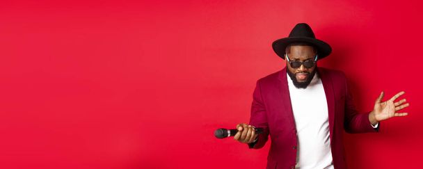 Passionate black male singer performing against red background, singing into microphone, wearing party outfit, standing over red background. - 写真・画像