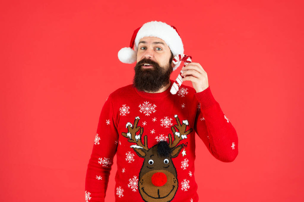 positive bearded guy in santa claus hat and sweater hold candy stick on red background, santa. - Фото, зображення