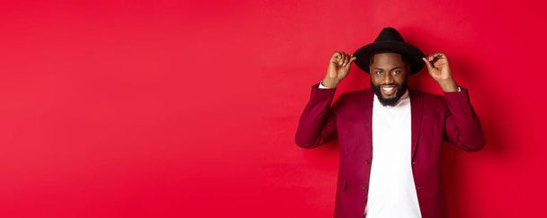 Мода и концепция вечеринки. Красивый африканский американец в шляпе и пиджаке, улыбающийся, уверенный в себе, стоящий на красном фоне. - Фото, изображение