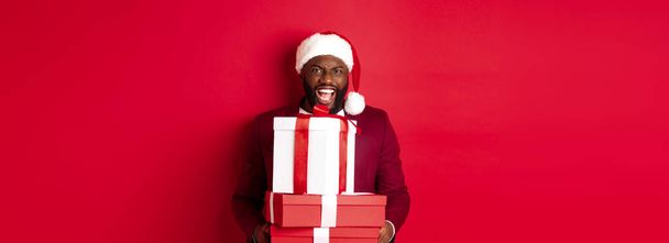 Christmas, New Year and shopping concept. Happy Black man in santa hat and blazer holding xmas presents, bring gifts and smiling, standing against red background. - Photo, Image