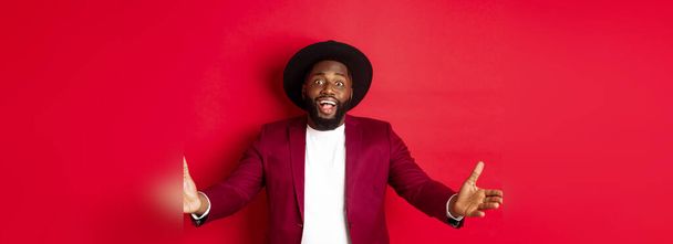 Fashion and party concept. Pleased handsome Black man spread hands in welcome gesture, reaching to receive something, looking amazed, standing over red background. - Photo, Image