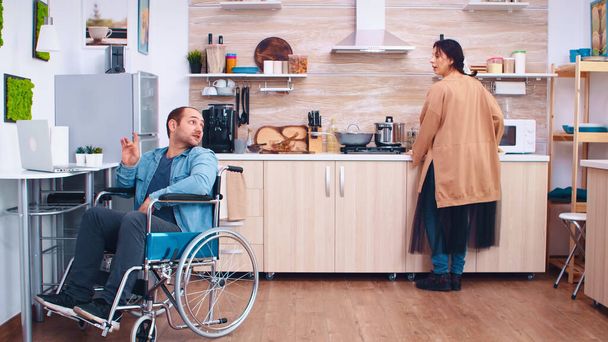 Ευτυχισμένος ανάπηρος σε αναπηρική καρέκλα χρησιμοποιώντας φορητό υπολογιστή στην κουζίνα. Ο άνθρωπος που μιλάει σε βιντεοδιάσκεψη. Εταιρικός άνδρας με αναπηρία αναπηρία αναπηρία αναπηρία δυσκολίες εργασίας μετά το ατύχημα έχοντας - Φωτογραφία, εικόνα