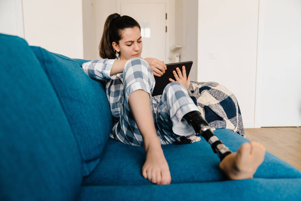 Young woman with prosthesis using tablet computer while resting on couch at home - Φωτογραφία, εικόνα