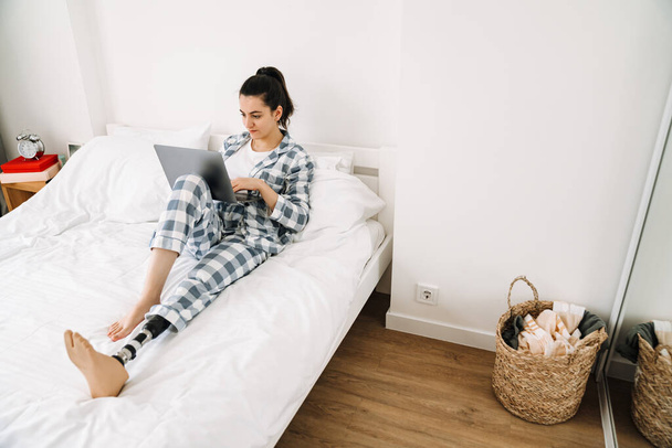 Young woman with prosthesis using laptop while resting on bed at home - Foto, Bild
