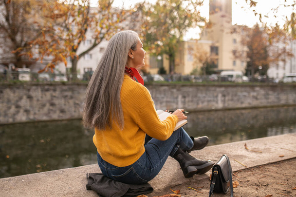 Künstler. Reife Frau sitzt am Flussufer und zeichnet in Album - Foto, Bild