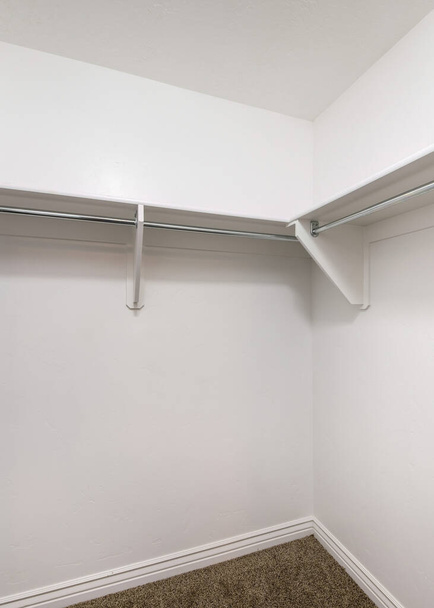 Vertikale weiße Regale mit Holzkonsolen und Metallstangen in einem begehbaren Kleiderschrank. Innenraum eines begehbaren Kleiderschranks mit weißen Wänden und Regalen über dem braunen Teppichboden. - Foto, Bild