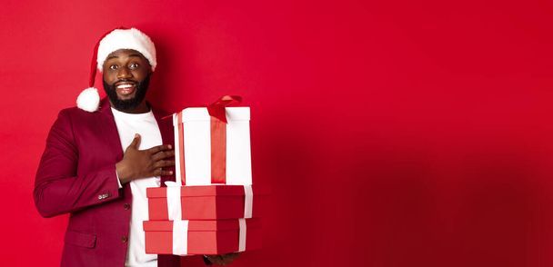 Karácsony, újév és vásárlási koncepció. Boldog fekete férfi kap karácsonyi ajándékokat, azt mondja, köszönöm, és mosolygós hálás, álló Mikulás kalap ellen piros háttér. - Fotó, kép