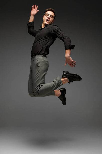 High jumps. Happy young man, student, business in black classic style shirt and grey trousers running, jumping isolated over grey background. Modern office style, emotions, active lifestyle - Fotó, kép