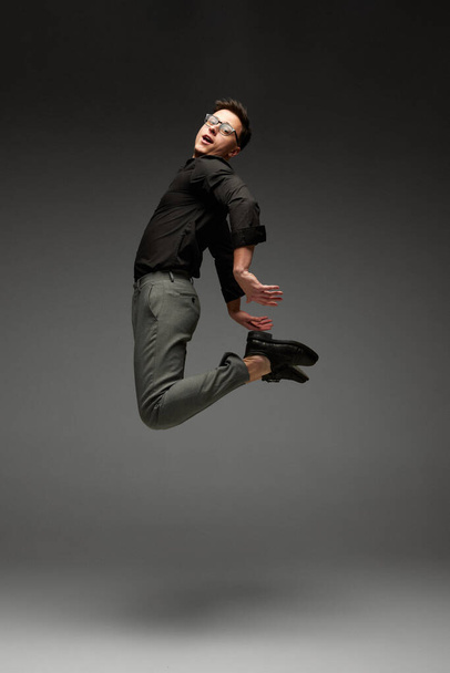 Freedom. Happy young man, student, business in black classic style shirt and grey trousers running, jumping isolated over grey background. Modern office style, emotions, active lifestyle - Foto, afbeelding