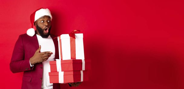 Karácsony, újév és vásárlási koncepció. Meglepett fekete férfi nézi karácsonyi ajándékok csodálkozva, visel Mikulás kalap, álló ajándékokkal szemben piros háttér. - Fotó, kép