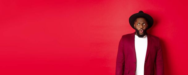 Fashion and party concept. Shocked and displeased black man staring at something unpleasant, looking upper left corner and frowning, standing against red background. - Photo, Image