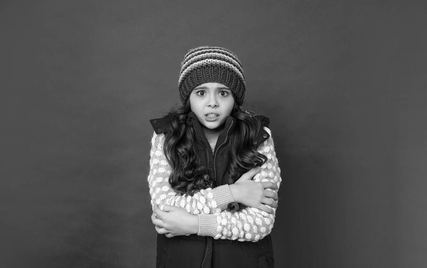 teen girl feel cold in knitted winter hat and down vest on red background, cold season. - Foto, Imagen