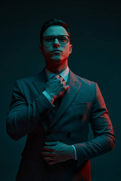 Fashionable young businessman in classic style suit adjusting neck tie isolated over dark background in neon light. Concept of business, fashion, style, modern lisfestyle. - Foto, Imagen