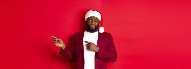 Christmas, party and holidays concept. Upset african american man looking gloomy, close eyes and sighing, pointing fingers right, standing in santa hat against red background. - Фото, изображение