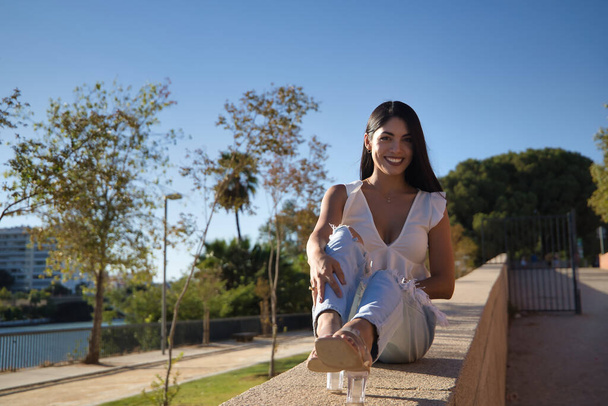 South American woman, young and beautiful, brunette, with white shirt and jeans, sitting on a stone wall, smiling and happy. Concept beauty, sunlight, happiness, smile. - Foto, Bild