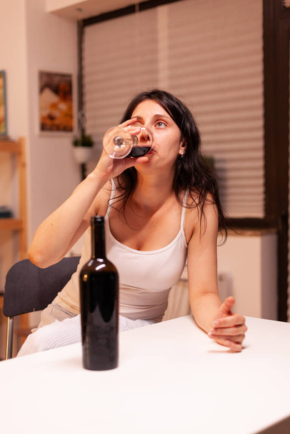 Vrouw met levensproblemen die veel alcohol drinkt aan de keukentafel. Ongelukkig persoon ziekte en angst zich uitgeput voelen met alcoholisme problemen. - Foto, afbeelding