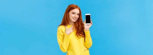 Impressed and fascinated redhead sassy woman touch heart and flirty gazing camera showing sexy guy photo she met dating app, holding smartphone with display facing camera, blue background. - 写真・画像