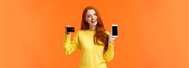 Shopping, Online- und Finanzierungskonzept. fröhlich junge rothaarige Frau eröffnet Bankkonto, hält Smartphone und Kreditkarte, lächelt, empfehlen Online-Shopping-Anwendung. - Foto, Bild