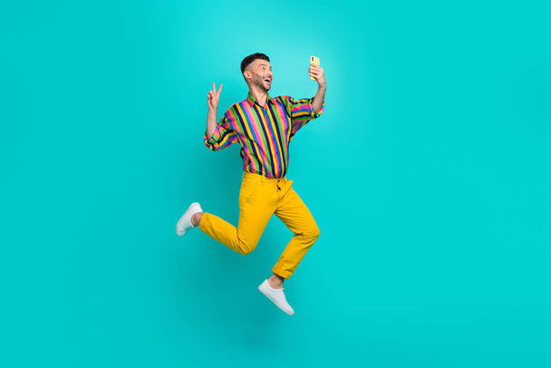 Full body photo of funny jumping air guy wear striped shirt showing v-sign selfie with smartphone hello followers isolated on cyan color background. - Fotografie, Obrázek