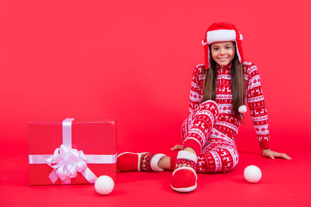new year. positive santa teen girl with new year present sit in studio. teen girl with new year santa present isolated on red background. - Foto, Imagen