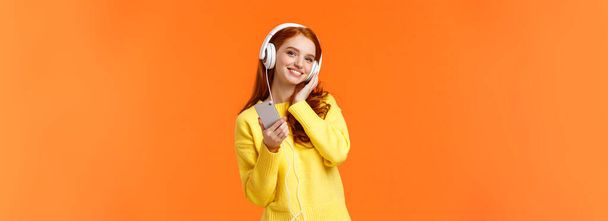 Waist-up portret vrolijke mooie jonge vrouw met rood haar luisteren muziek in hoofdtelefoon, kantelen hoofd en glimlachen tevreden, holding smartphone, kies lied voor zachte rustige avond, oranje achtergrond. - Foto, afbeelding