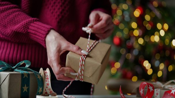 Burgonya rengi süveterli bir kadın, içinde hediyeler olan bir sürü kutuyu güzel kağıt ve kurdelelerle paketliyor, bir Noel ağacının arka planında fiyonklar. Noel, yeni yıl tatili, hediye paketi.. - Fotoğraf, Görsel