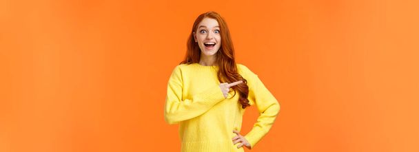 Happy, upbeat and dreamy cute redhead woman got chance travel abroad, telling about awesome company, promoting product on sale, stare fascinated camera smiling and pointing left, orange background, - Photo, Image